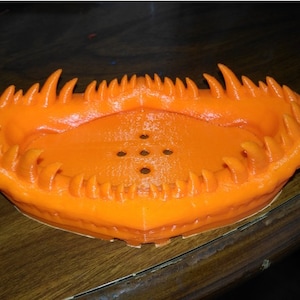 May include: Orange plastic dish with a jagged, toothy rim. The dish has a series of small holes in the bottom.
