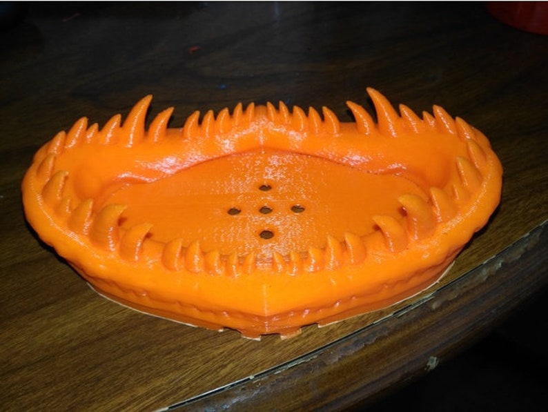 May include: Orange plastic dish with a jagged, toothy rim. The dish has a series of small holes in the bottom.