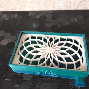 Puede incluir: Un jabonera impresa en 3D de color azul verdoso y verde con un dise&ntilde;o geom&eacute;trico blanco. La jabonera tiene un dise&ntilde;o de flor de loto en la parte inferior.