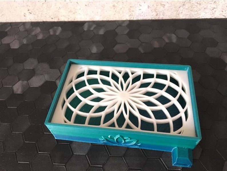 Puede incluir: Un jabonera impresa en 3D de color azul verdoso y verde con un dise&ntilde;o geom&eacute;trico blanco. La jabonera tiene un dise&ntilde;o de flor de loto en la parte inferior.