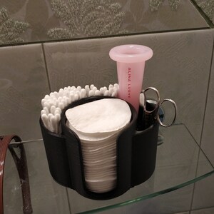 May include: A black plastic organizer with compartments for cotton swabs, cotton rounds, and other bathroom essentials. A pink cup with the text "Alina Lurye" is in the organizer. A pair of silver tweezers is also in the organizer.