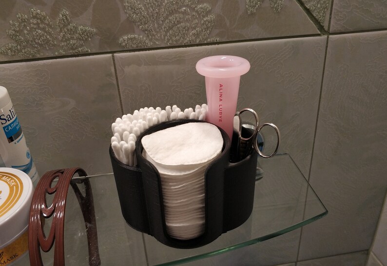 May include: A black plastic organizer with compartments for cotton swabs, cotton rounds, and other bathroom essentials. A pink cup with the text "Alina Lurye" is in the organizer. A pair of silver tweezers is also in the organizer.