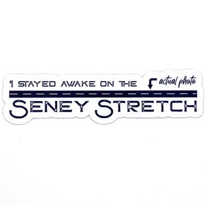 May include: A white sticker with navy blue text. The text reads "I STAYED AWAKE ON THE actual photo SENEY STRETCH" with a road graphic. The sticker is rectangular with rounded corners.