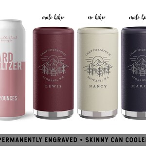 May include: Three personalized skinny can coolers with a mountain scene and the text "Camp Fitzpatrick, Spokane, WA". The coolers are in burgundy, beige, and navy blue. The text "Lewis", "Nancy", and "Marcus" are engraved on the coolers.