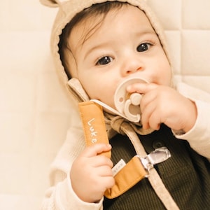 May include: A baby wearing a beige knitted hat with ear flaps, a green and beige outfit, and a pacifier. The hat has a brown strap with the name "Luke" embroidered on it.