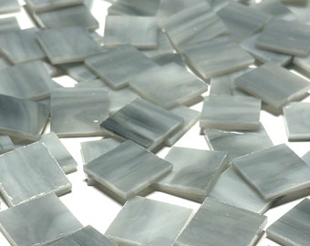 May include: A pile of small, square, gray glass tiles with a white marbled pattern. The tiles are scattered on a white surface.