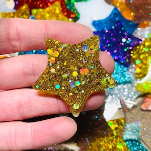 May include: A gold glitter star-shaped embellishment with iridescent specks. The star is held in a hand, with other colorful glitter stars in the background. This decorative item is suitable for crafts and DIY projects.