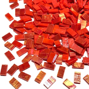May include: A pile of small, rectangular, red glass mosaic tiles. The tiles are scattered across a white surface, with varying shades of red and some with a reflective, iridescent quality. These tiles are suitable for craft projects.