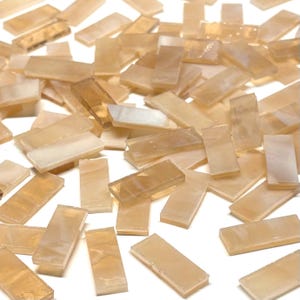 Puede incluir: Un conjunto de pequeñas teselas rectangulares de mosaico en tonos beige y crema. Las teselas están dispersas, creando una superficie texturizada. La cualidad translúcida de las teselas permite el paso de la luz, realzando sus sutiles variaciones de color.