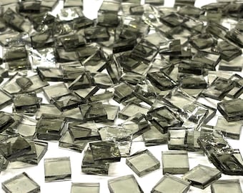 May include: A collection of small, square, gray glass tiles scattered on a white surface. The tiles are translucent and reflect light, creating a shimmering effect. Ideal for mosaic art projects or decorative applications.