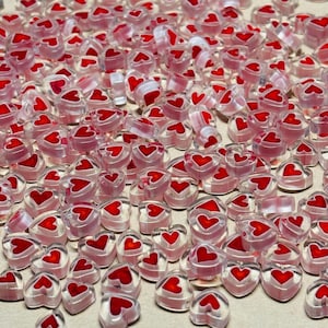 May include: A large quantity of small, clear heart-shaped beads with red centers. The beads are scattered on a white surface.