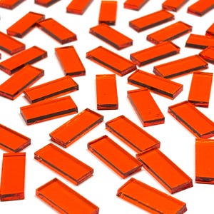 May include: A collection of rectangular, translucent orange glass tiles scattered on a white surface. The tiles vary slightly in size and shape, with a dark line visible on the edges. These tiles are suitable for mosaic art projects.