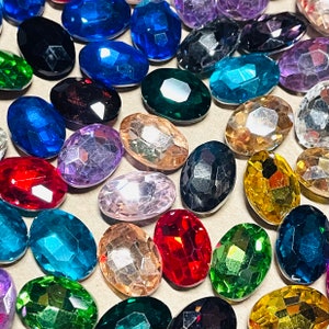May include: A collection of oval-shaped, faceted acrylic gemstones in a variety of vibrant colors including red, blue, green, yellow, purple, black, and clear. Each gem features a polished, multi-faceted surface that reflects light.