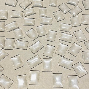 Peut inclure: Un grand nombre de cabochons en verre transparents et rectangulaires éparpillés sur une surface brune. Les cabochons sont tous de la même taille et de la même forme, et ils ont un dessus légèrement bombé.