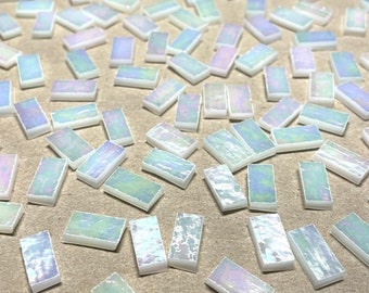 Puede incluir: Una colección de azulejos rectangulares blancos iridiscentes, esparcidos sobre una superficie marrón. Los azulejos tienen un brillo sutil y son perfectos para proyectos de manualidades o decoración del hogar.