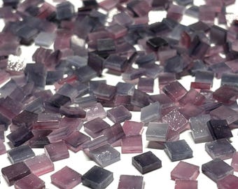 Puede incluir: Una gran cantidad de pequeñas baldosas cuadradas de vidrio iridiscente en tonos de morado y gris. Las baldosas están esparcidas sobre una superficie blanca.