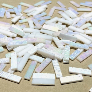 Pode incluir: Uma coleção de pequenas tesselas retangulares para mosaicos. As tesselas são principalmente brancas com detalhes iridescentes e perolados. As tesselas estão espalhadas sobre uma superfície, mostrando suas formas e variações de cor. Ideal para projetos de artesanato.