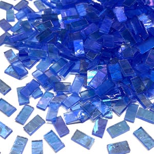 May include: A scattering of small, rectangular, blue glass mosaic tiles. The tiles are a vibrant shade of blue, with a slightly iridescent quality. The tiles are scattered across a white surface, creating a textured and colorful display. Ideal for craft projects.