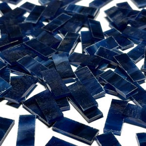 May include: A collection of small, rectangular, dark blue glass tiles scattered on a white surface. The tiles have a slightly translucent quality, with variations in the blue hue. Ideal for mosaic projects or decorative crafts.