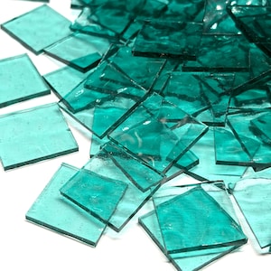 May include: A collection of small, square-shaped glass tiles in a translucent teal color. Each tile features a smooth, flat surface with slightly irregular edges, creating a semi-transparent appearance when layered together in a scattered pile.