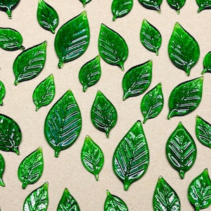 May include: A collection of glossy, emerald green glass leaves with detailed veining. The leaves vary in size and are arranged on a neutral, light brown surface. The glass has a reflective quality, enhancing the vibrant green color.