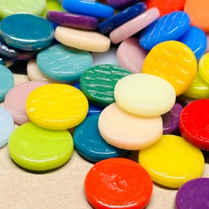 May include: A collection of colorful, flat, round glass mosaic tiles. The tiles are in various shades of blue, green, yellow, red, purple, and pink. Some tiles have a textured, striped pattern. These decorative tiles are suitable for crafts and mosaic projects.