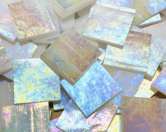 Puede incluir: Un montón de azulejos de mosaico de vidrio blanco iridiscente con una sutil variación de color azul y marrón. Los azulejos son cuadrados y tienen un acabado liso y brillante.