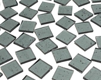 May include: A collection of small, square, gray glass tiles scattered on a white surface. Each tile has a slightly reflective surface and dark gray edges. The tiles are evenly distributed, creating a mosaic effect.