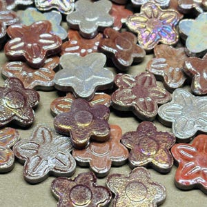 May include: A collection of flower-shaped ceramic beads in various colors, including silver, bronze, and reddish-brown. The beads have a glossy finish and are scattered across a surface, suggesting their use in jewelry making or crafts.
