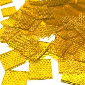 May include: A collection of small, square, translucent yellow glass tiles. Each tile features a distinct raised diamond-shaped grid pattern on its surface, creating a textured appearance. The tiles are scattered in a pile, showing their vibrant color.