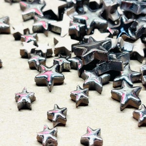 May include: A collection of small, metallic star-shaped embellishments. The stars have a silver finish with a pinkish hue on the edges, scattered across a neutral surface. These decorative elements are ideal for crafts and jewelry making.