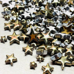 May include: A collection of metallic star-shaped embellishments in gold and silver tones. The stars vary in size and are scattered across a light-colored surface. The shiny stars are likely for crafting or decorative purposes.