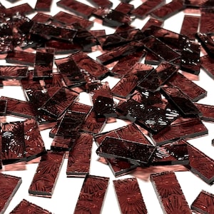 May include: A pile of small, rectangular, dark red glass tiles with a textured surface. The tiles are scattered on a white background.