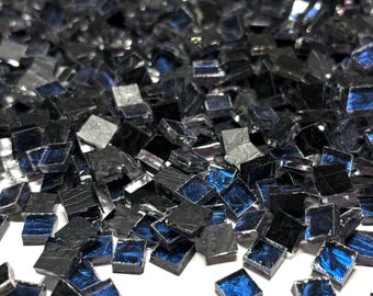 Könnte beinhalten: Ein Haufen kleiner, quadratischer, irisierender Glasfliesen in Schwarz- und Blautönen. Die Fliesen sind auf einer weißen Oberfläche verstreut.