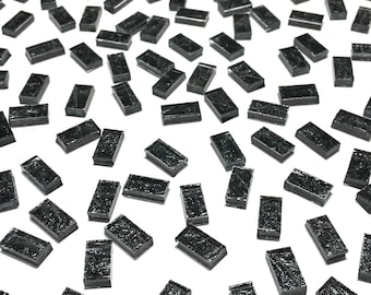 Könnte beinhalten: Eine Streuung kleiner, rechteckiger, dunkelgrauer Glasfliesen. Die Fliesen sind auf einer weißen Oberfläche verstreut, jede mit einem leicht strukturierten Aussehen. Der Gesamteffekt ist eine Ansammlung kleiner, reflektierender, dunkelgrauer Rechtecke.