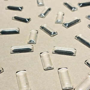 May include: A collection of small, clear, rectangular glass cabochons with rounded edges and a smooth, domed top surface. These transparent, elongated pieces are scattered across the frame, each reflecting light with a polished, glossy finish.