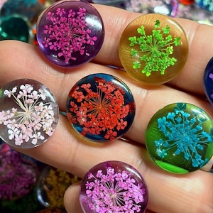 May include: A collection of circular resin cabochons, each featuring a pressed, dried flower in a distinct color. The set includes vibrant shades of pink, green, light pink, red, and blue, all encased in clear, smooth, polished resin domes.