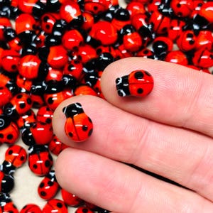 May include: A collection of small, red ladybug beads with black heads and spots. Two beads are held between fingers, showcasing the details of the insect-shaped glass beads. The background is filled with more of the same ladybug beads.