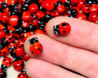 Può includere: Primo piano di perline rosse a forma di coccinella con teste e punti neri. Due perline sono tenute tra le dita, mostrando i dettagli delle perline di vetro a forma di insetto. Lo sfondo è pieno di altre perline a coccinella.