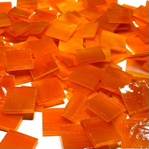 May include: A collection of small, square, orange glass tiles. The tiles are translucent with a glossy finish. They are scattered on a white surface, creating a mosaic effect. The colour is a warm, inviting orange.