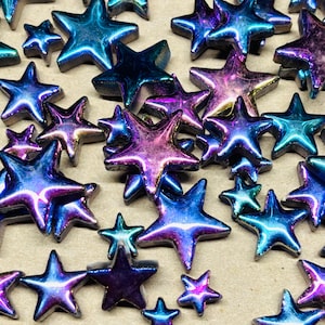 May include: An assortment of iridescent star-shaped embellishments in various sizes. The stars display a color-shifting effect, with hues of blue, purple, and pink. These decorative elements are scattered across a neutral surface, creating a celestial theme.