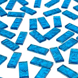 May include: A collection of rectangular blue glass tiles scattered across a white surface. Each tile features a raised, textured sunburst or floral pattern on its top surface, with a translucent finish and smooth, straight edges throughout.