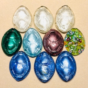 May include: A collection of nine oval-shaped glass beads in various colors. The beads include clear, green, light blue, purple, and blue, with a textured, shell-like pattern. One bead features a colorful floral design.
