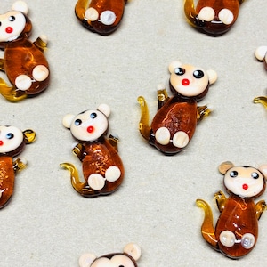 May include: A collection of small, handcrafted glass monkey figurines. Each monkey has a brown body, white belly and ears, and a red mouth. The monkeys have yellow tails and arms. The background is a light gray color.