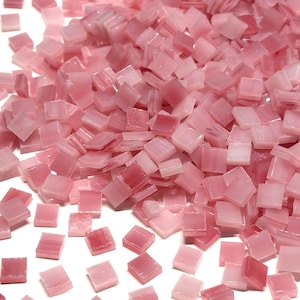 May include: A collection of small, square-shaped glass mosaic tiles in various shades of light and medium pink. The tiles have a smooth, glossy finish and are scattered across the frame, displaying slight variations in colour and translucency.