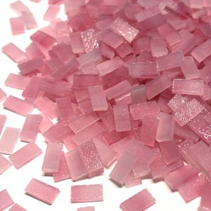 May include: A pile of small, rectangular, pink glass mosaic tiles. The tiles vary in shades of pink, from light to medium, and appear translucent. The tiles are scattered, creating a textured surface. Ideal for craft projects, these tiles offer a delicate, pastel aesthetic.