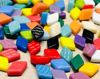 Può includere: Una collezione di tessere di mosaico di vetro a forma di diamante colorate in vari colori, tra cui giallo, arancione, blu, rosso e viola. Alcune tessere hanno superfici strutturate. Le tessere sono sparse, creando un'esposizione vibrante e artistica.