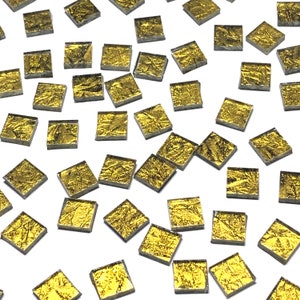 May include: A collection of small, square, gold-colored glass tiles with a textured surface. The tiles are scattered on a white background.