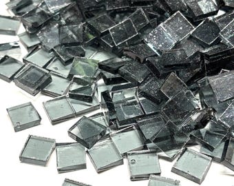 May include: A pile of small, square, dark gray glass tiles. The tiles have a reflective, slightly textured surface and are scattered across a white background. The tiles are likely for mosaic art or craft projects.