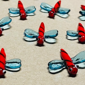 Op de afbeelding: Een verzameling van acht glazen libellenfiguren met rood-zwarte gestreepte lichamen en doorschijnende blauwe vleugels.
